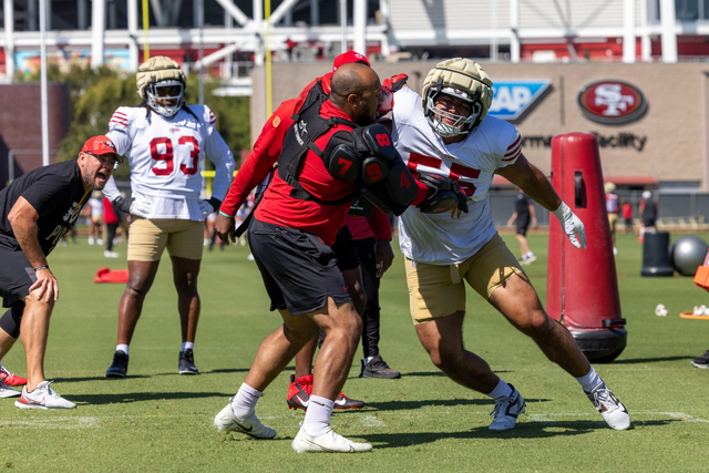 A football player wearing a white jersey and gold shorts pushes against a coach in a red padded vest during a practice drill.